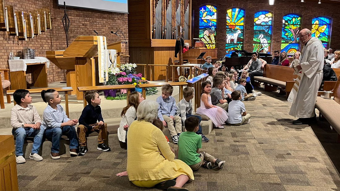 Children gathered at the front of Trinity's sanctuary beneath its stained glass windows
